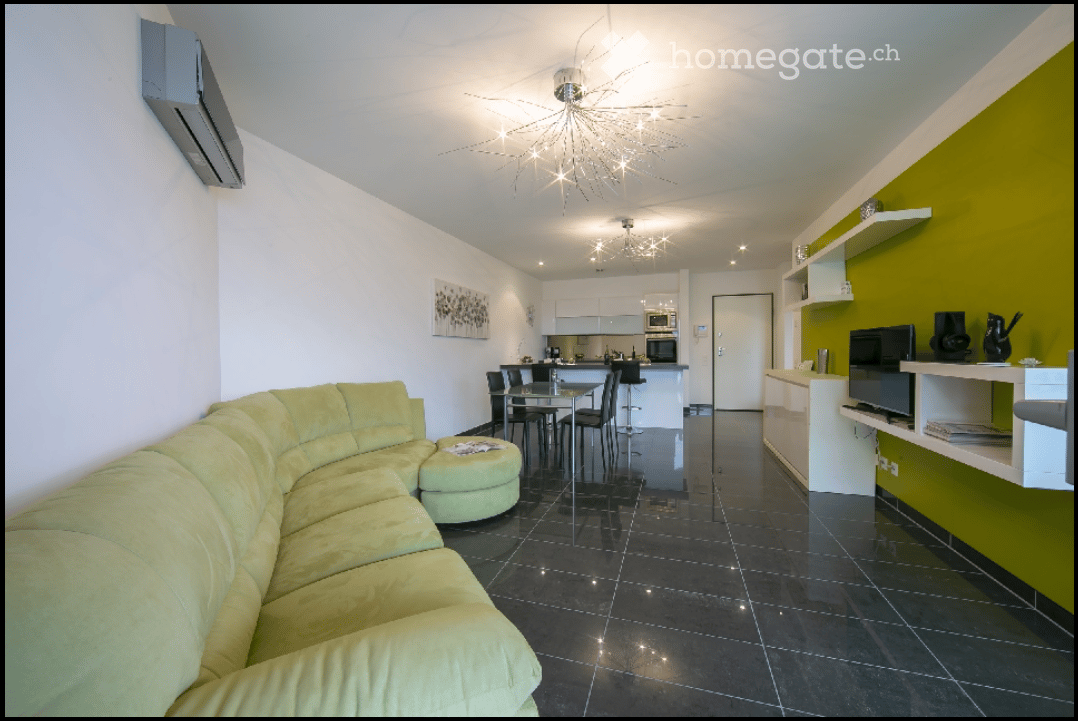 Modern living room with a green sofa, glass table, and a kitchen area in the background.