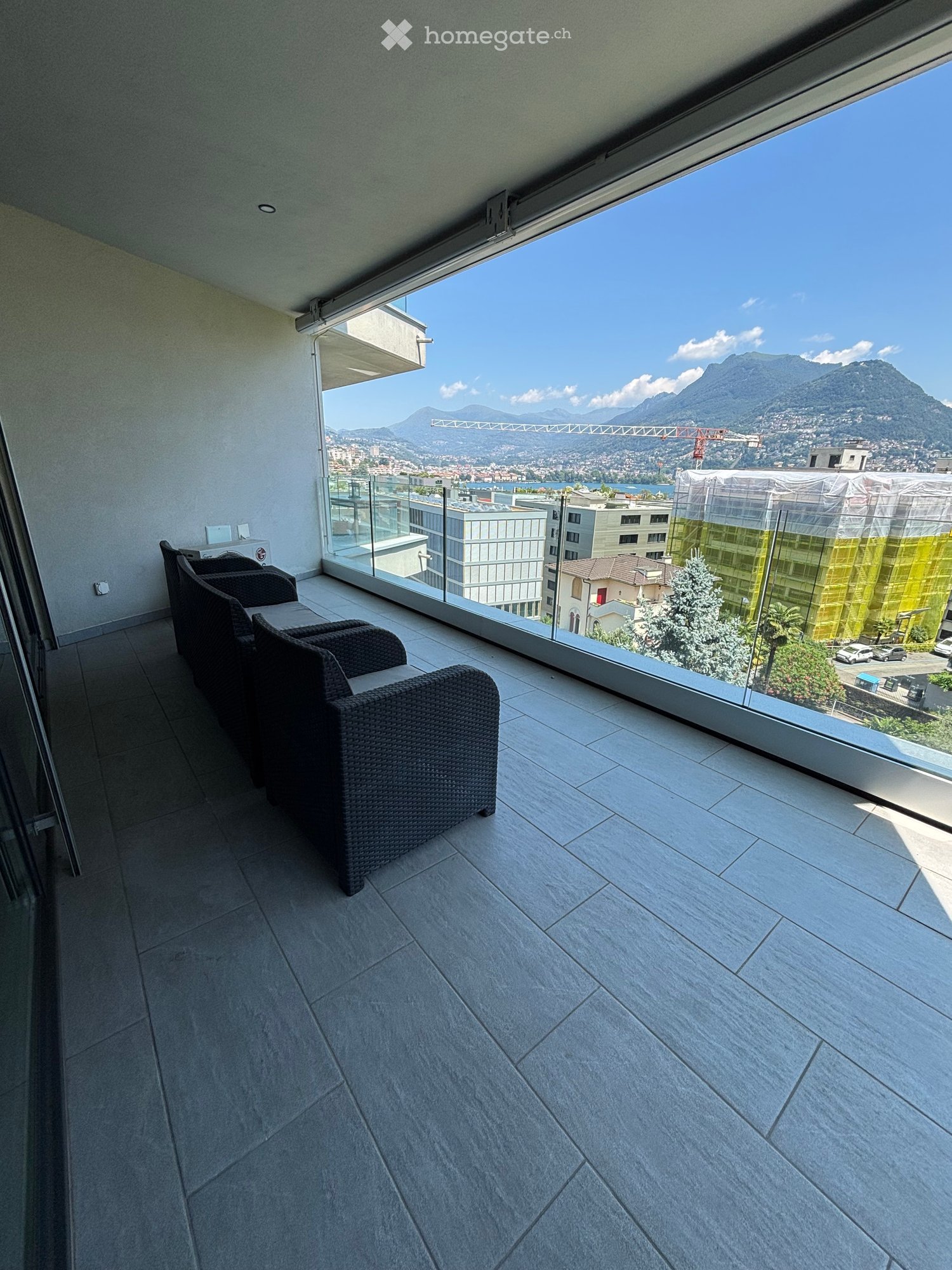 A balcony with a view of the city and mountains, featuring a tiled floor and a set of black chairs and a table.