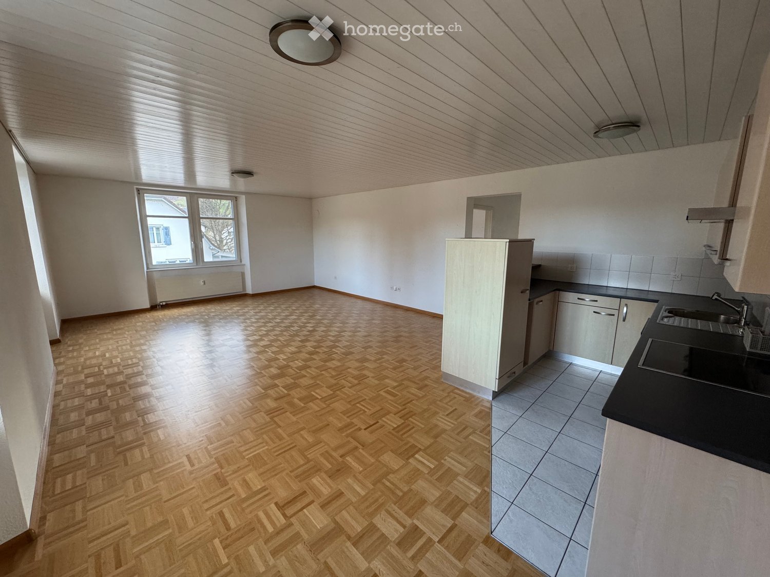 Empty room with wooden parquet flooring, white walls, and a modern kitchen area with a sink, cabinets, and a countertop.