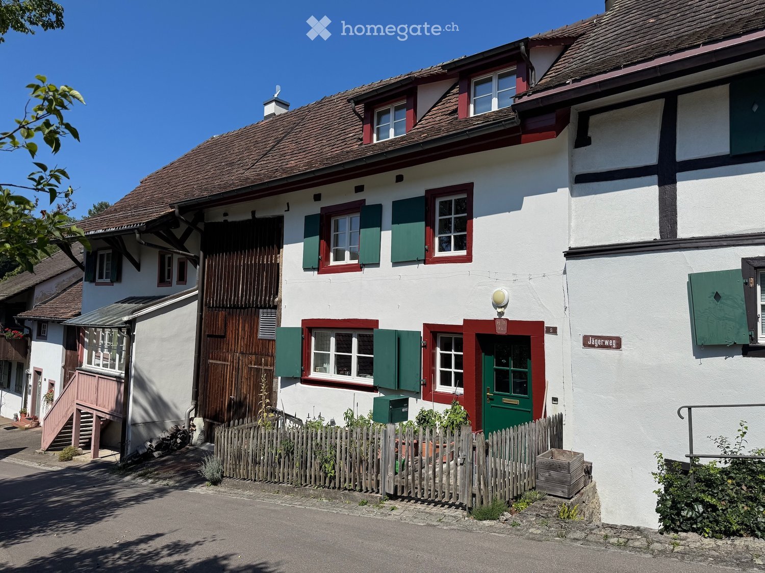 Two-story traditional house with white walls, red trim, green shutters, and a brown tiled roof. Features a wooden garage door, a small garden area with a wooden fence, and a sign reading "Jagerweg" above the entrance.