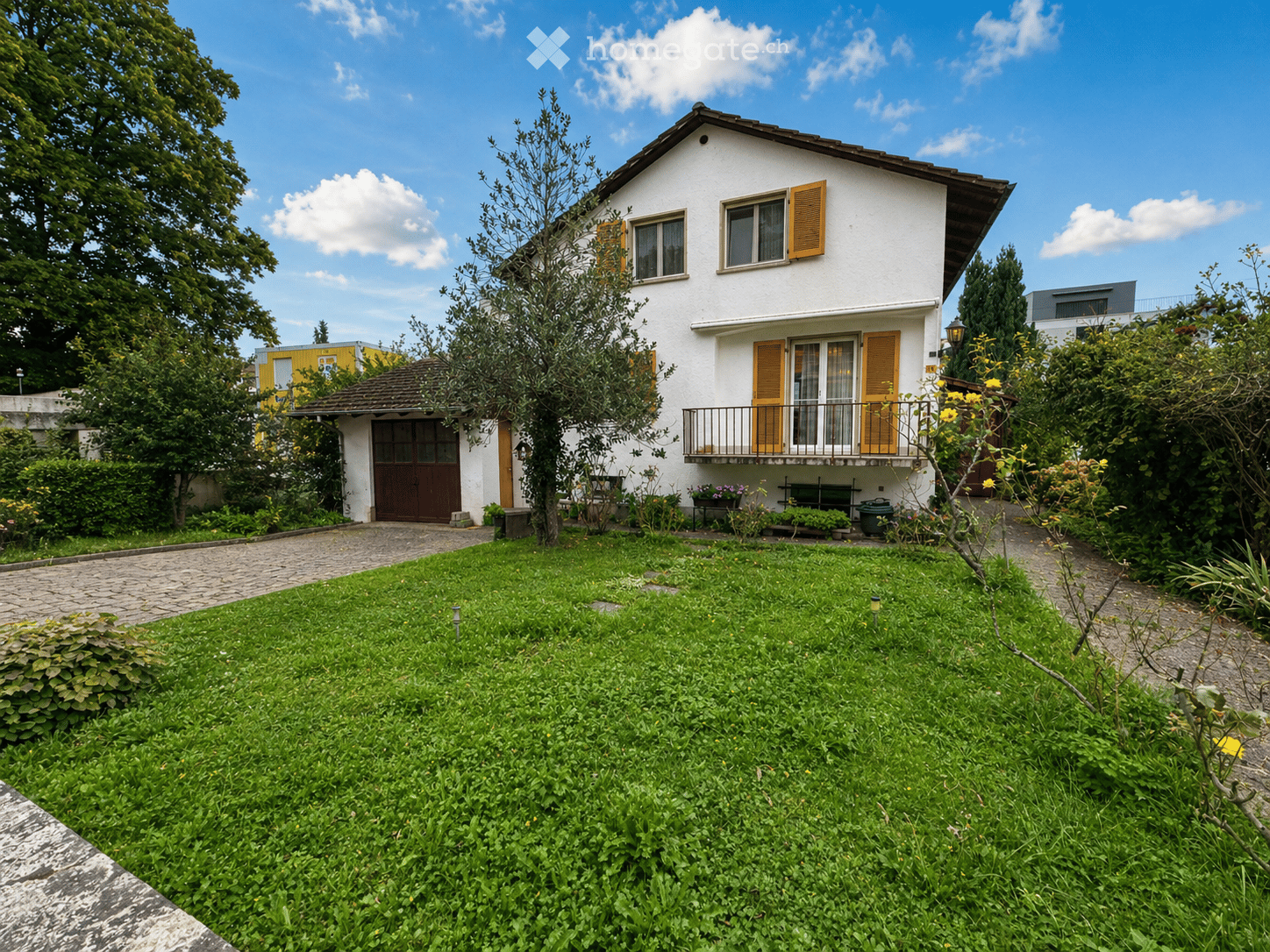 Two-story house with a brown roof, white walls, and yellow shutters. The house is surrounded by a well-maintained garden with various plants and trees. A driveway leads to the entrance, and there is a garage on the left side of the house.