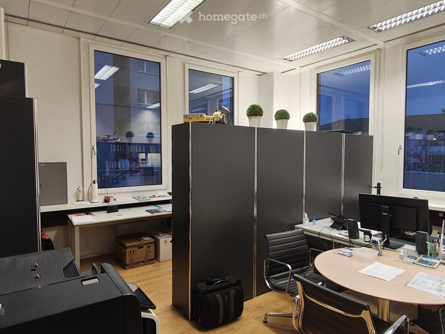 Wooden flooring, white walls, large windows, large black cabinets, desks, chairs, computer monitor, printer, table, boxes, potted plants