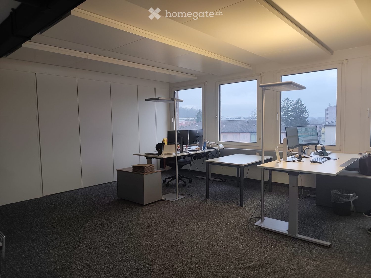 large room, grey flooring, large windows, two desks, two computer screens, adjustable height desks