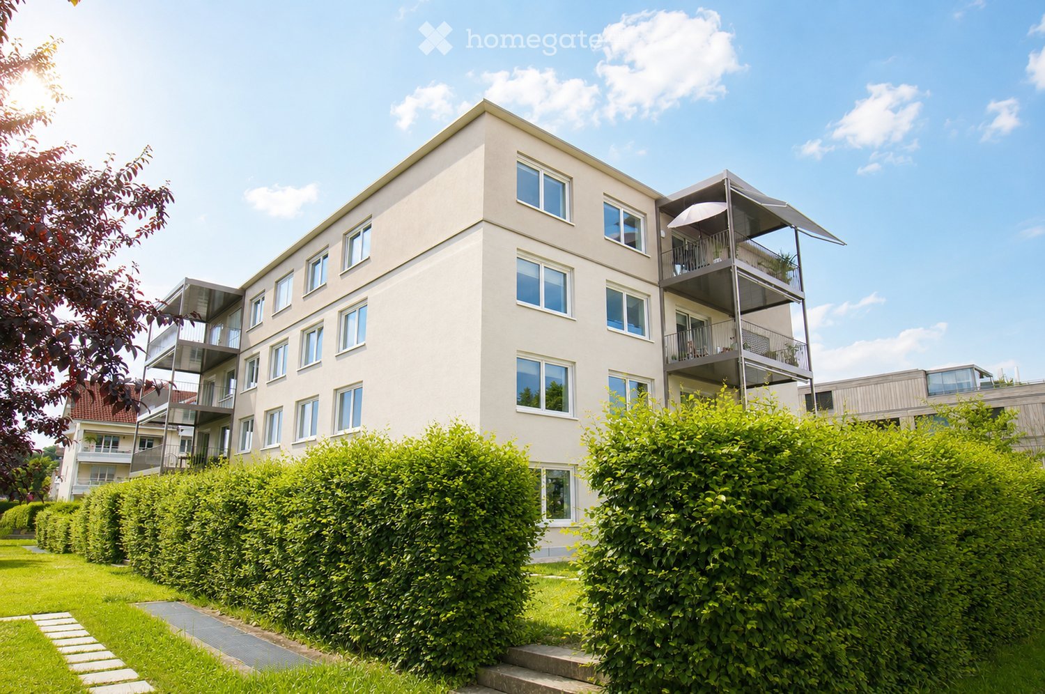Modern multi-story residential building with light beige facade, multiple windows, and balconies. Green hedges and lawn in front.