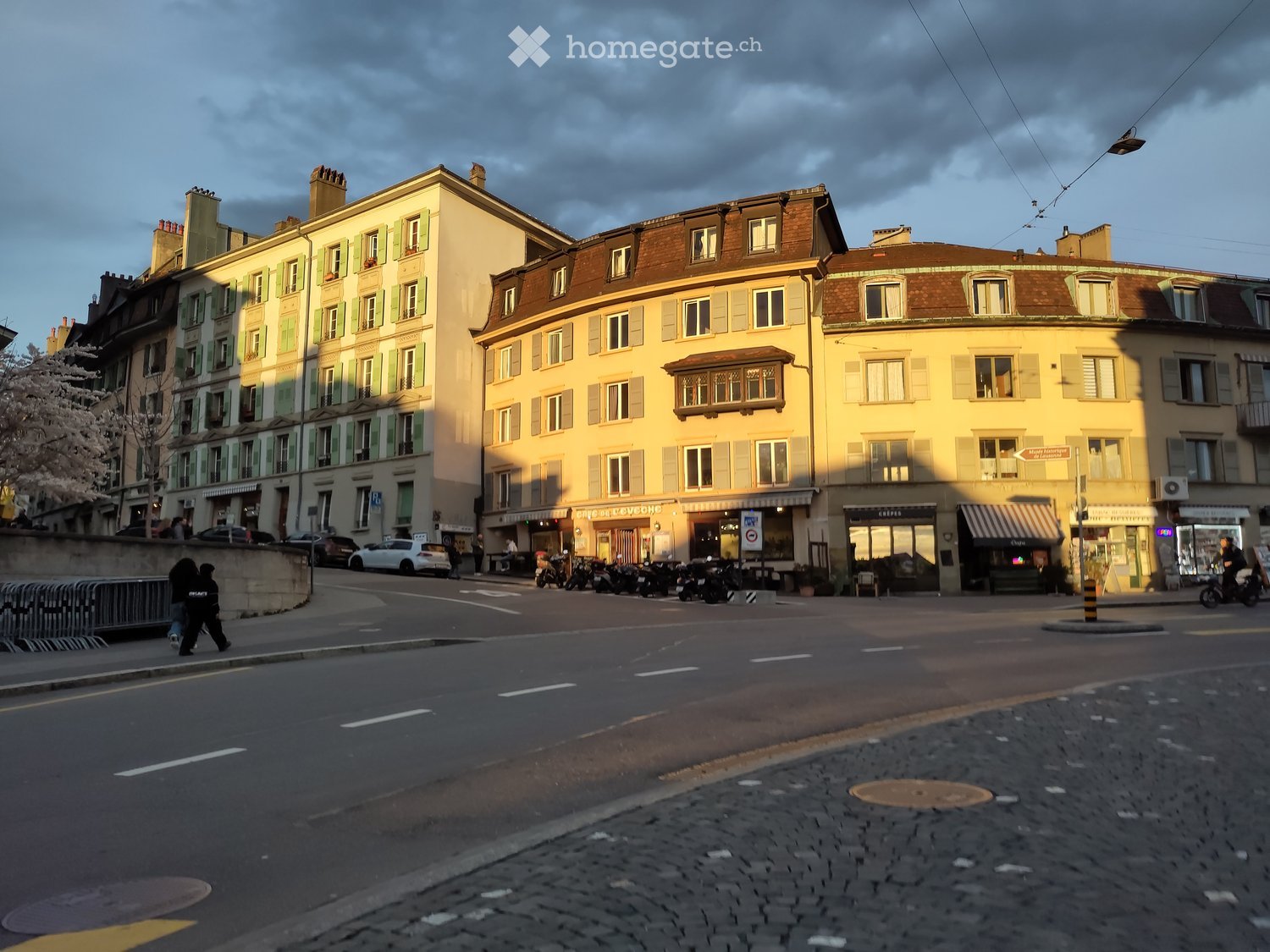 Building. A row of multi-story residential buildings with classic European architecture, featuring shutters, balconies, and storefronts at the ground level. The street is lined with parked motorcycles and a few cars, with pedestrians visible.