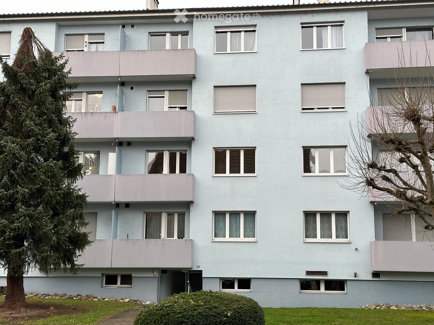 Four-story apartment building with light blue facade, multiple windows, and balconies. Green lawn and trees in front.