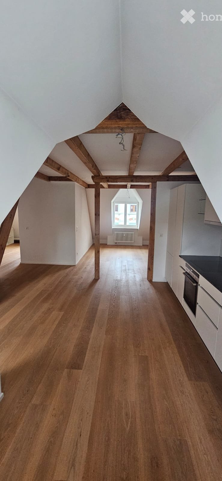 Empty attic space with wooden floors, exposed wooden beams, white walls, and a skylight window at the far end.