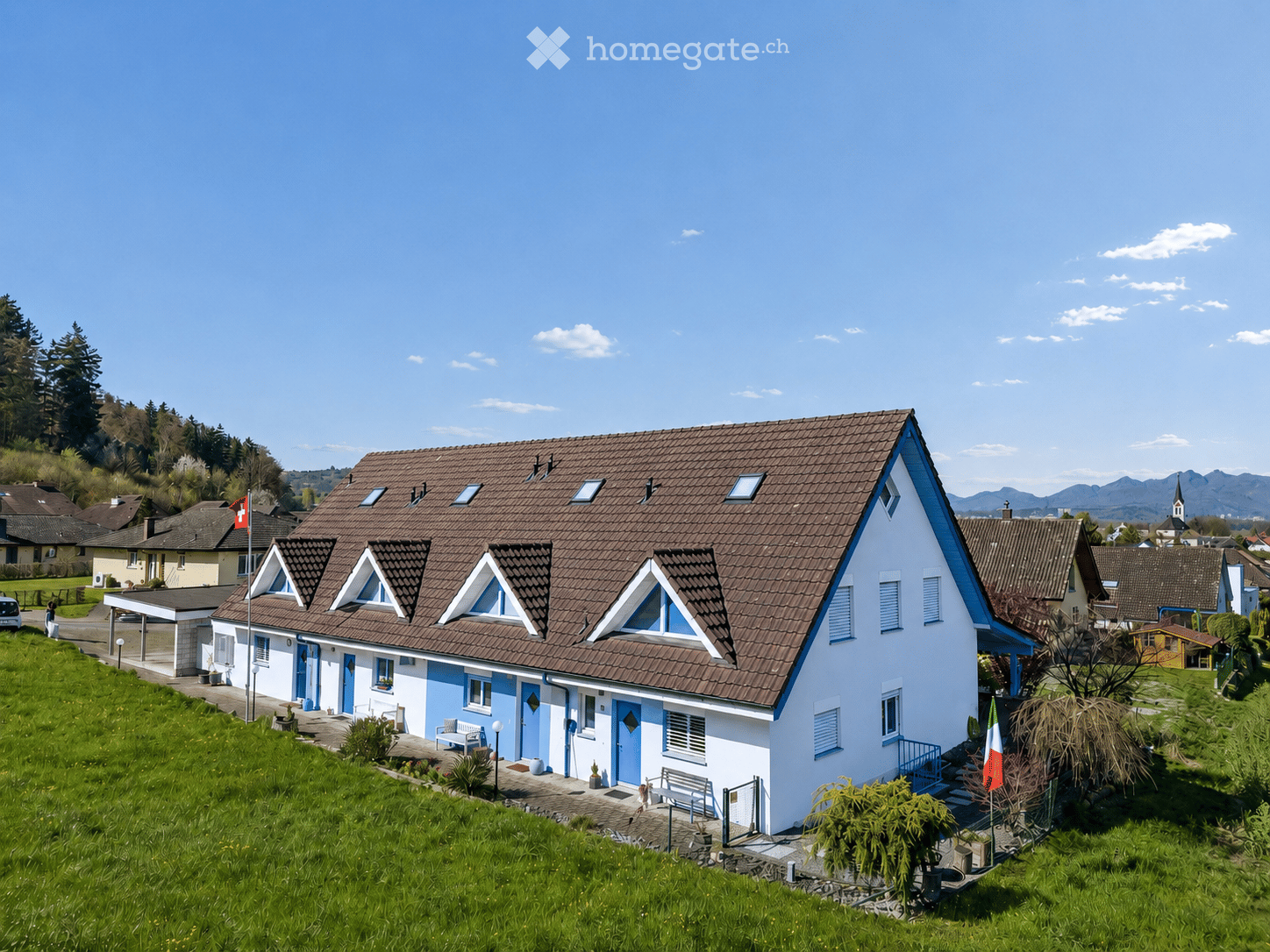 A two-story house with a brown tiled roof and blue trim, featuring multiple windows and dormer windows. The house is surrounded by a well-maintained lawn and garden, with neighboring houses visible in the background.