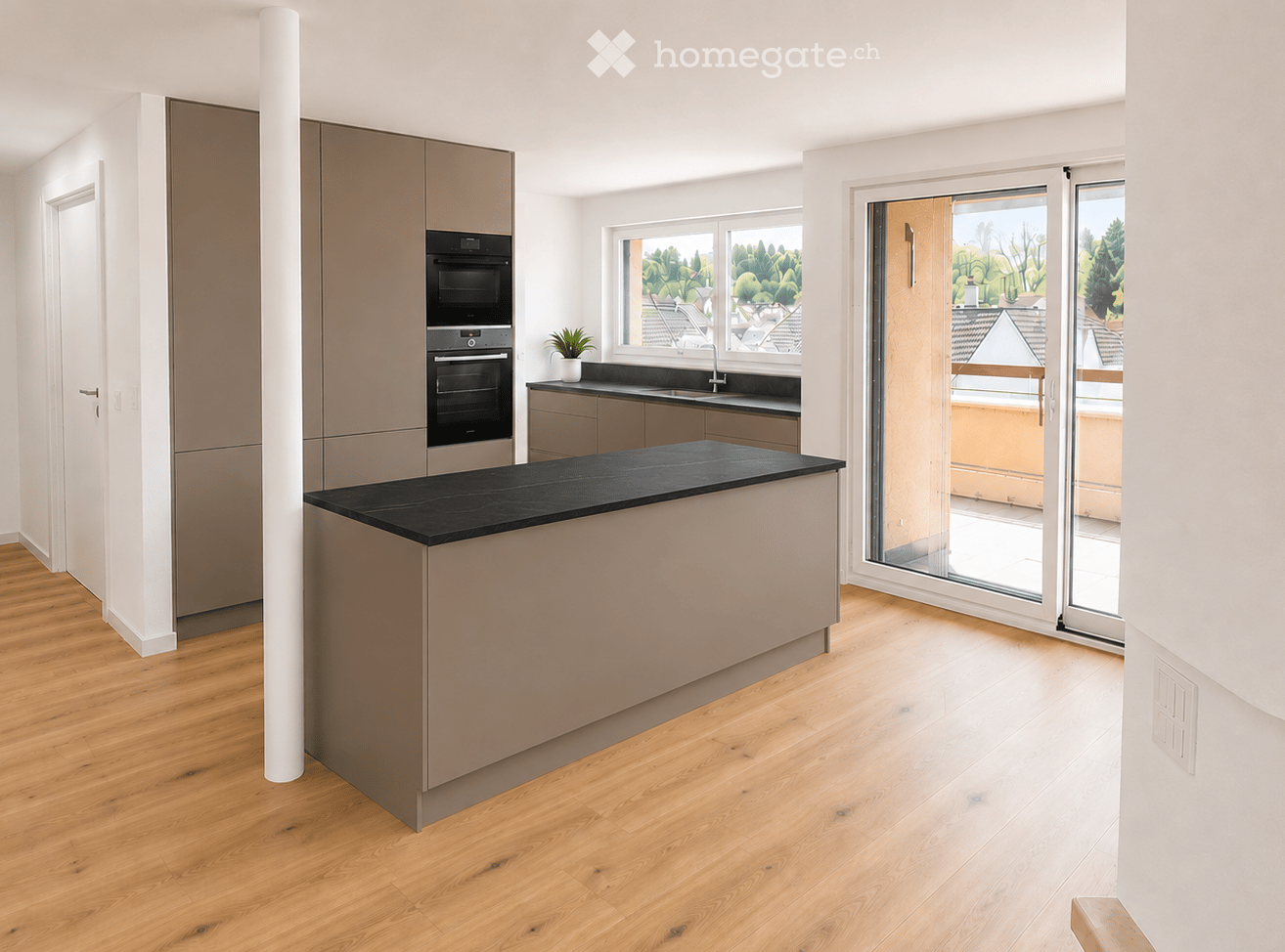 Modern kitchen with wooden flooring, sleek cabinets, built-in ovens, a central island with black countertop, and a view of greenery through large windows and sliding glass doors.