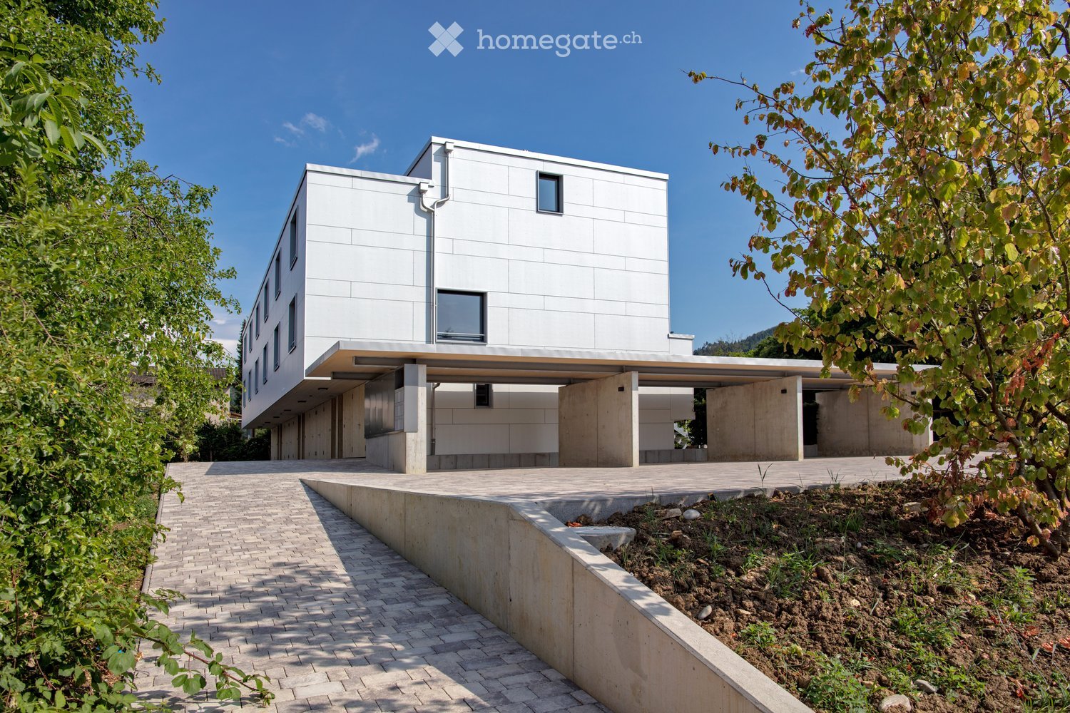 A modern, multi-story building with a white exterior and large windows. The building has a covered parking area and a paved walkway leading to the entrance. The surrounding area is landscaped with trees and greenery.