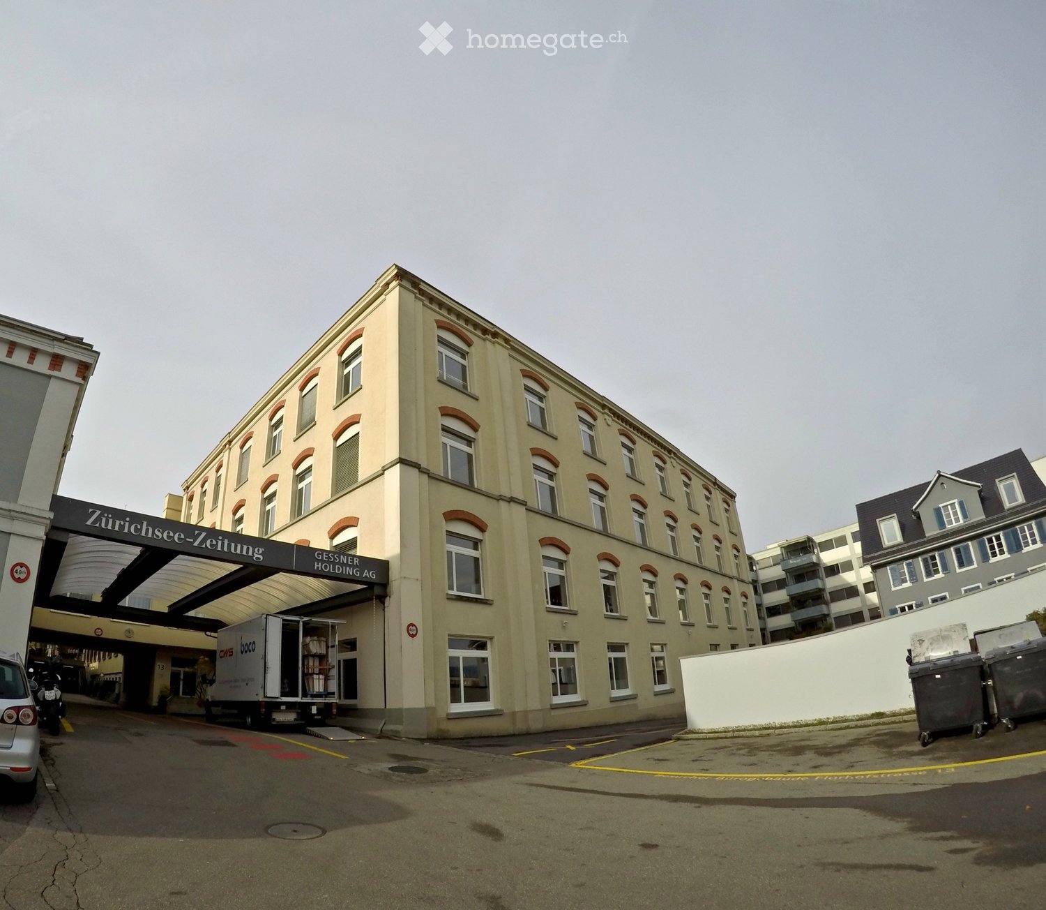 Commercial building with white walls and glass windows, several floors, name on the front entrance, cars and trash bins in the foreground