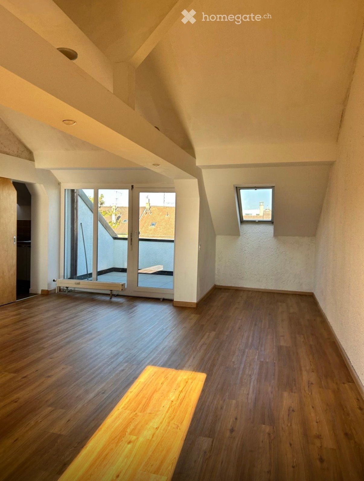 Empty attic room with wooden flooring, white walls, skylight window, and sliding glass door leading to a small balcony area.
