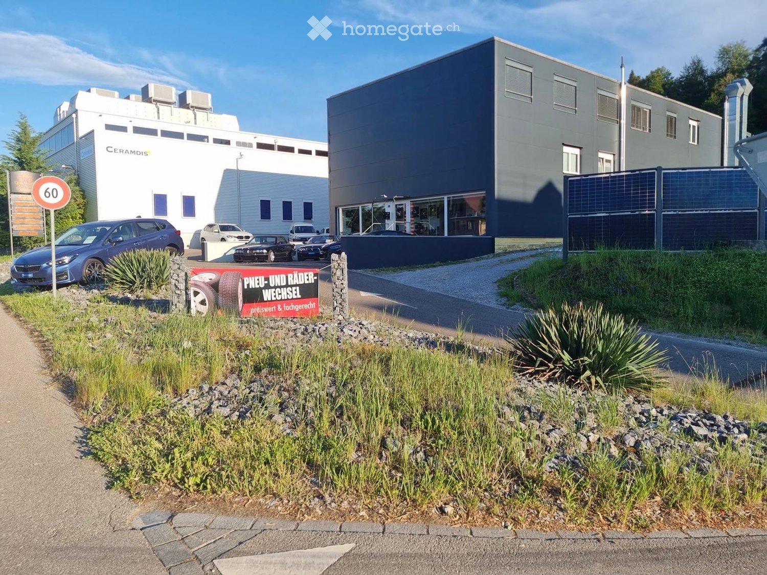 Building. Modern industrial building with a grey facade, large windows, and a sign reading "CERAMDIS" on the left. A parking area with several cars is visible in front of the building, along with a sign advertising tire and wheel exchange.