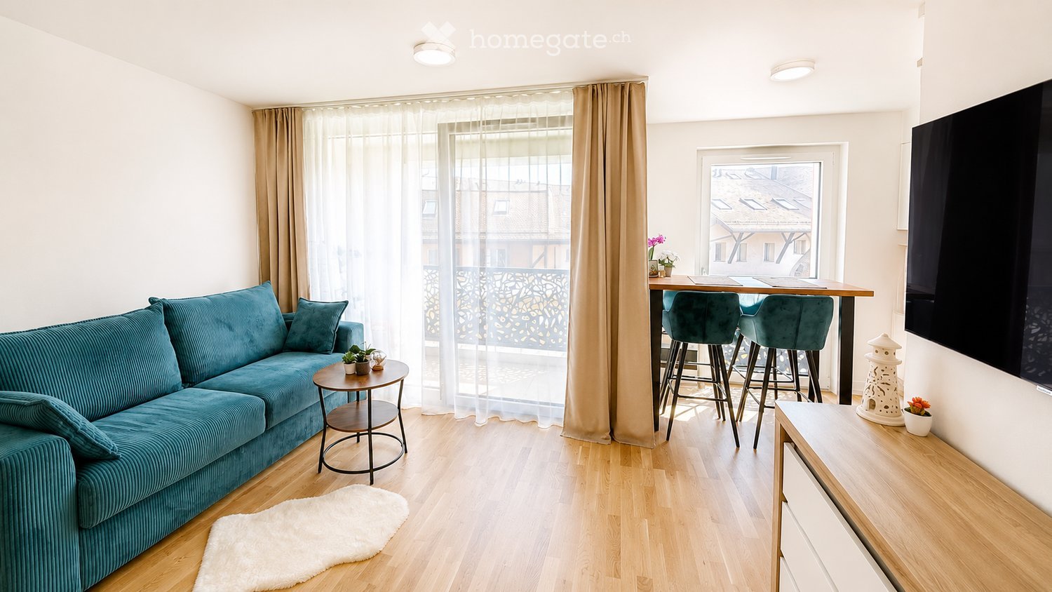 A modern living room with a teal sofa, coffee table, small side table, dining area with bar stools, and a flat-screen TV on the wall. The room features wooden flooring, large windows with sheer curtains, and a balcony view.