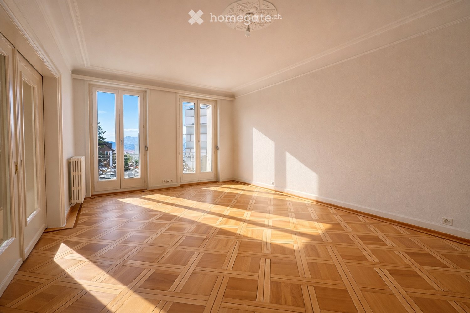 Empty room with wooden parquet flooring, large windows, and a radiator. Sunlight streams in, casting shadows on the floor.