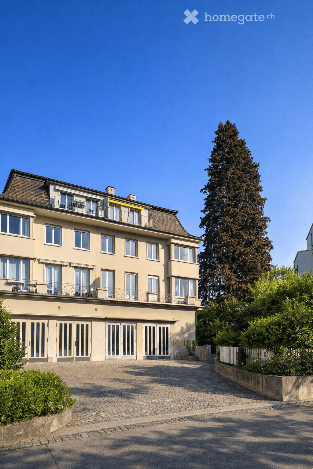 3 story building, beige exterior, multiple windows, balconies, driveway, garden