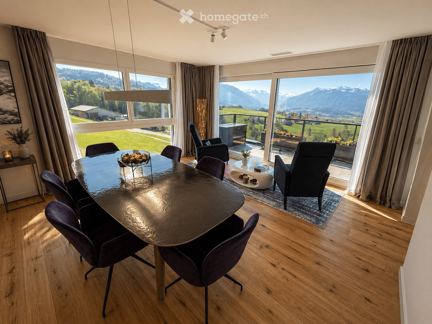 Modern dining area with a round glass table, purple chairs, and a view of the mountains through large windows. There is a small seating area with a sofa and coffee table near the balcony.