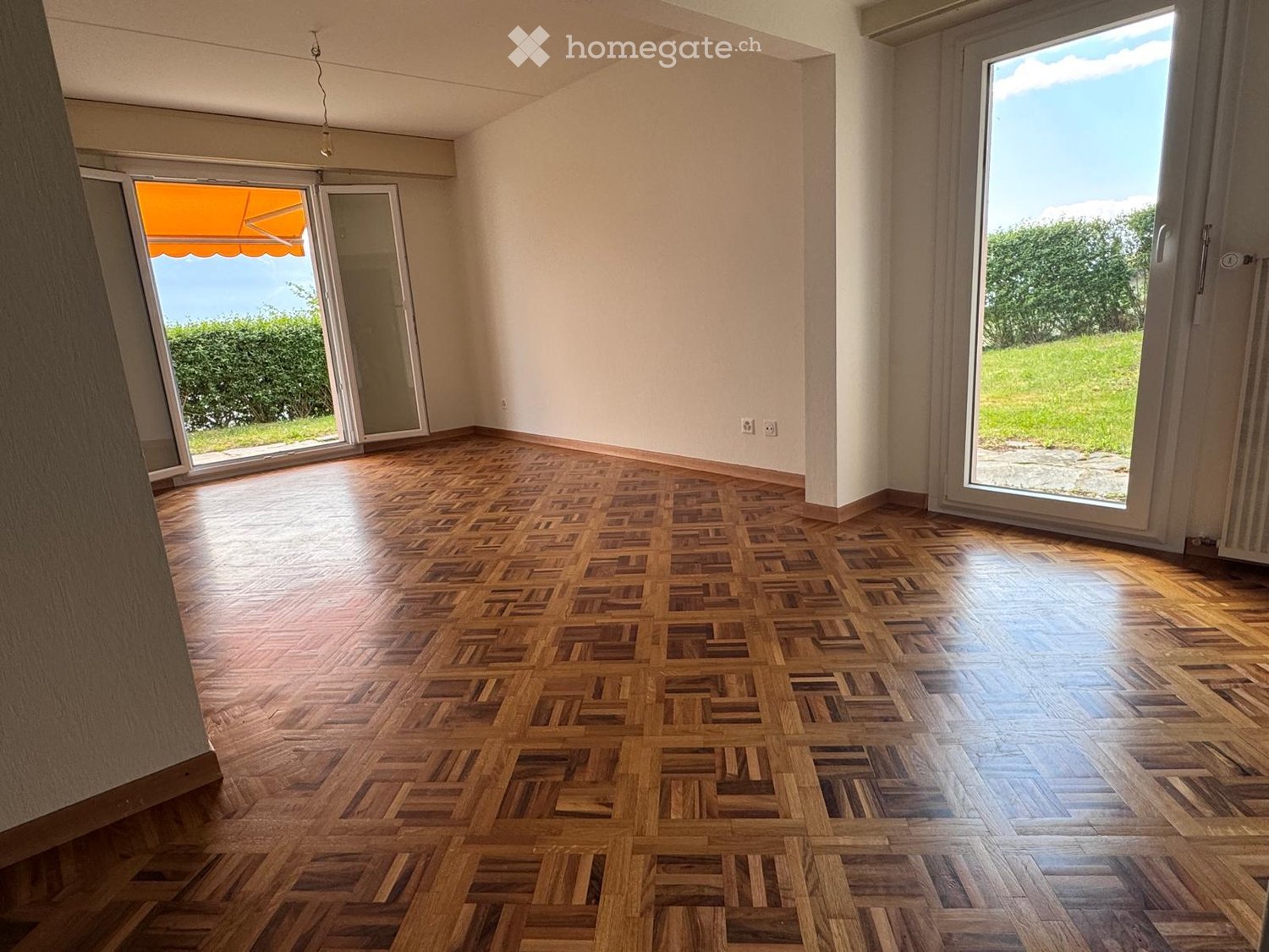 Empty room with wooden herringbone flooring, large sliding glass doors leading to a garden, and white walls.