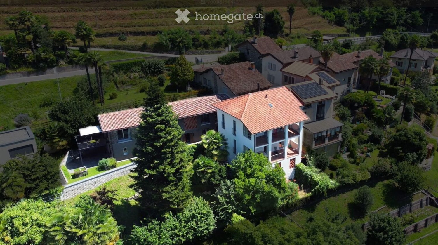 Aerial view of a residential area with multiple houses, surrounded by greenery and trees. The houses have red-tiled roofs and are set in a hilly landscape with a road running alongside.