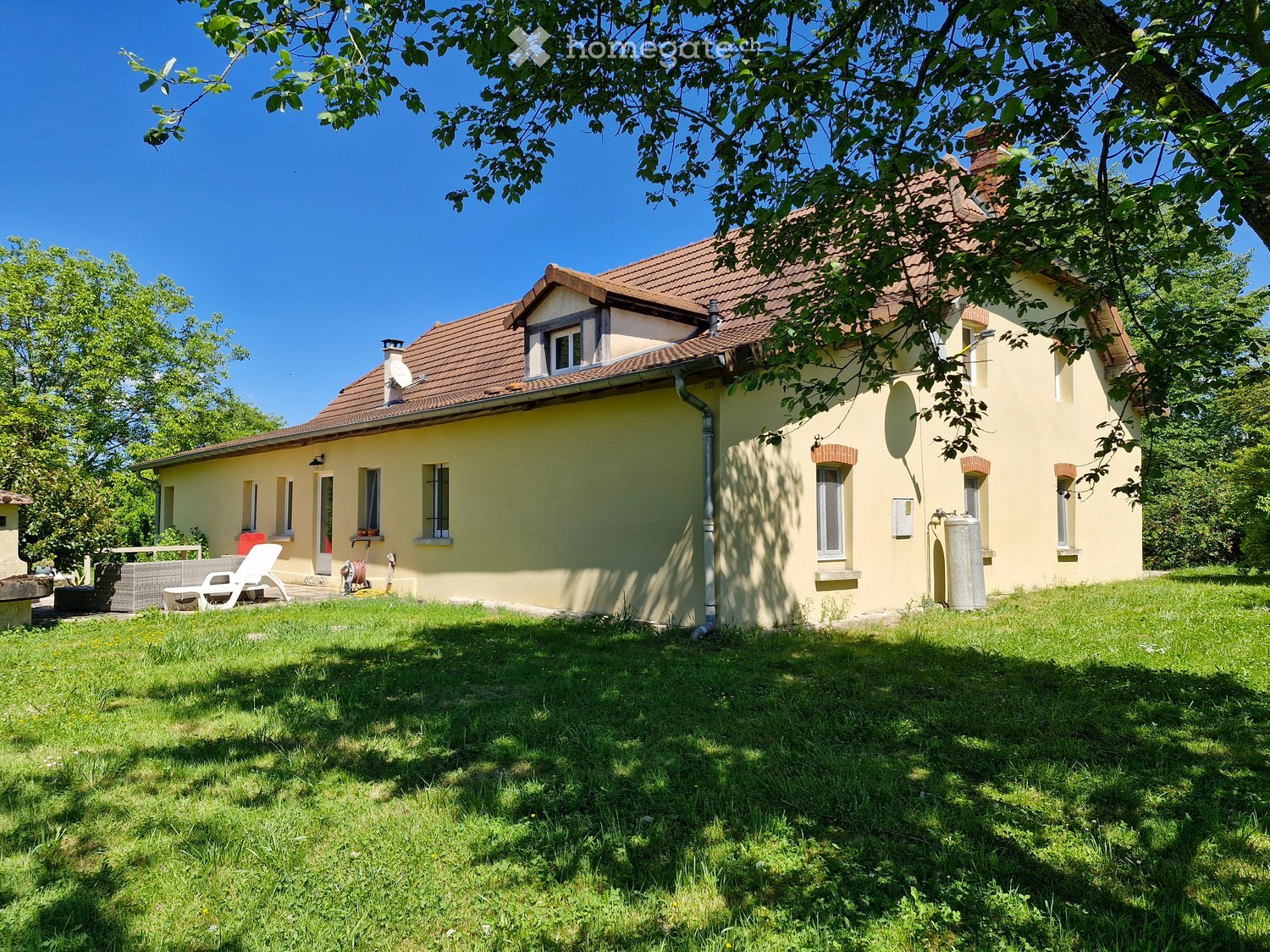 House. Single-story house with yellow walls, red-tiled roof, multiple windows, and a small garden area with outdoor furniture.