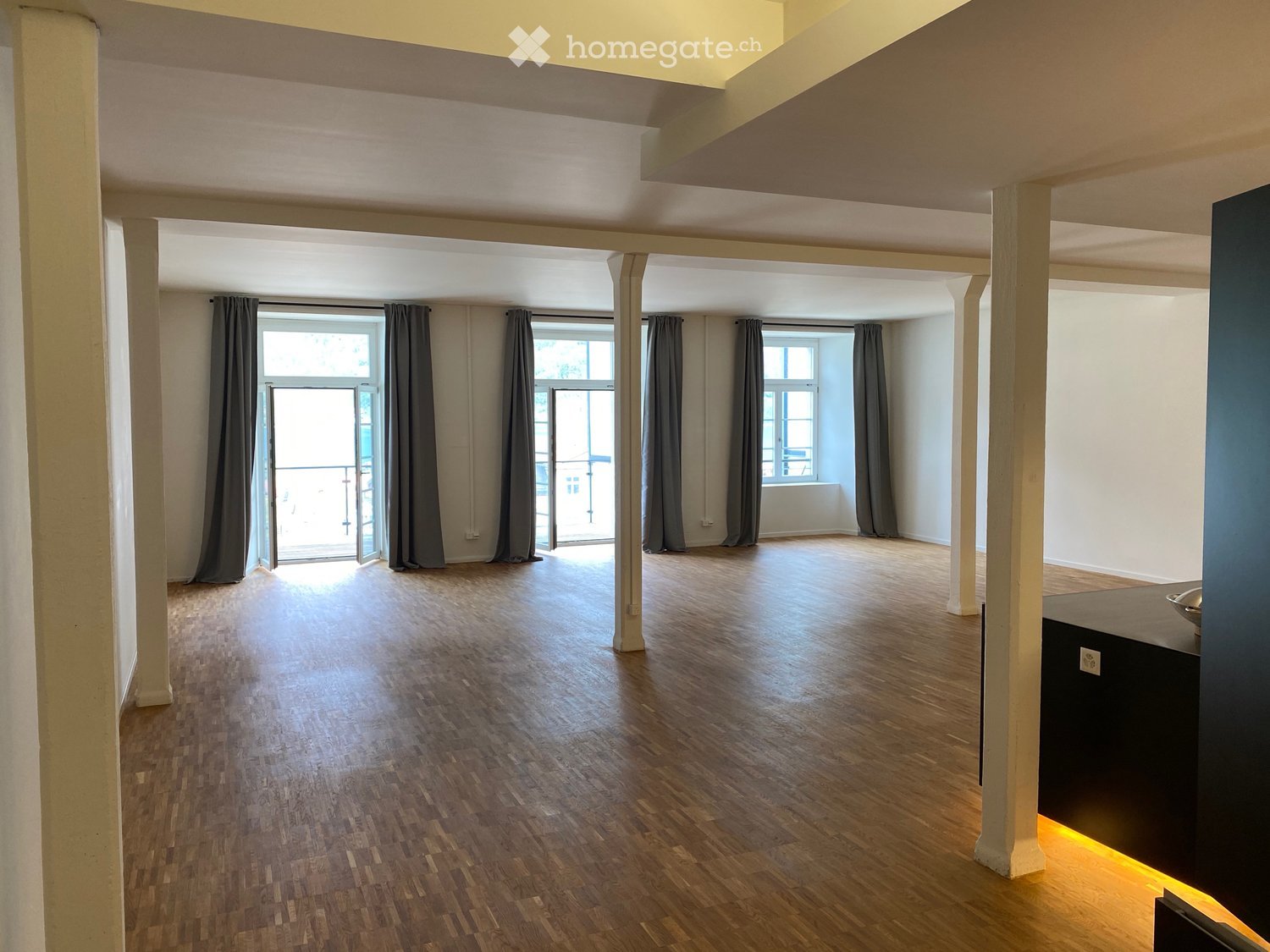 Empty room with wooden flooring, white walls, large windows with grey curtains, and a balcony accessible through glass doors.