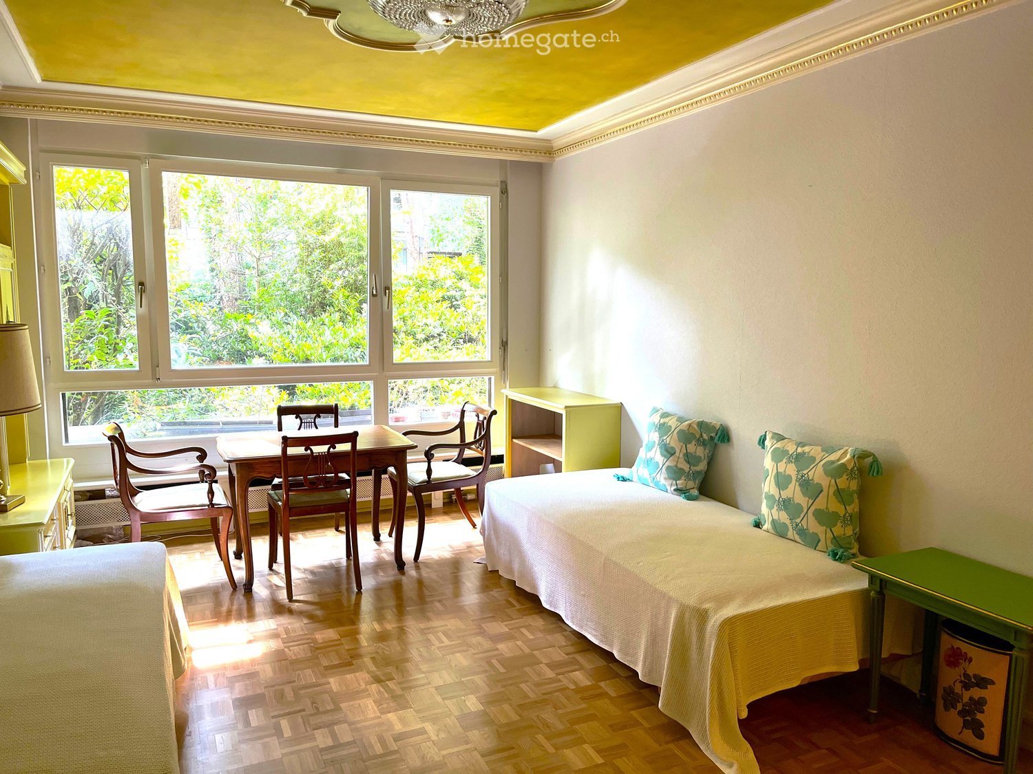 Room with hardwood floor, large windows, round dining table, chairs, couch, green desk, side tables, pillows, and lamp.