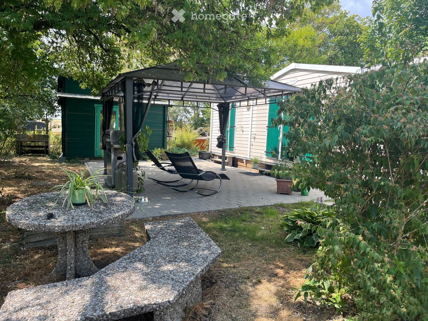 Outdoor patio area with a covered seating area, lounge chairs, a stone table, and surrounding greenery.