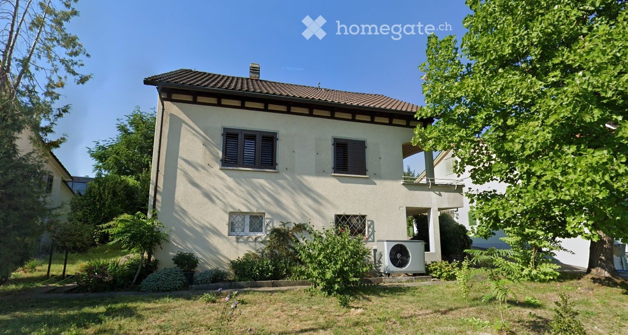 Two-story house with beige exterior, dark brown shutters, and a tiled roof. Features include a small balcony on the upper floor and an air conditioning unit on the ground floor.
