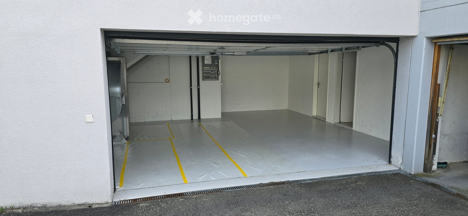Building. Empty garage with a concrete floor, white walls, and a large open door. Yellow lines are marked on the floor, and there is a small utility area visible on the left side.