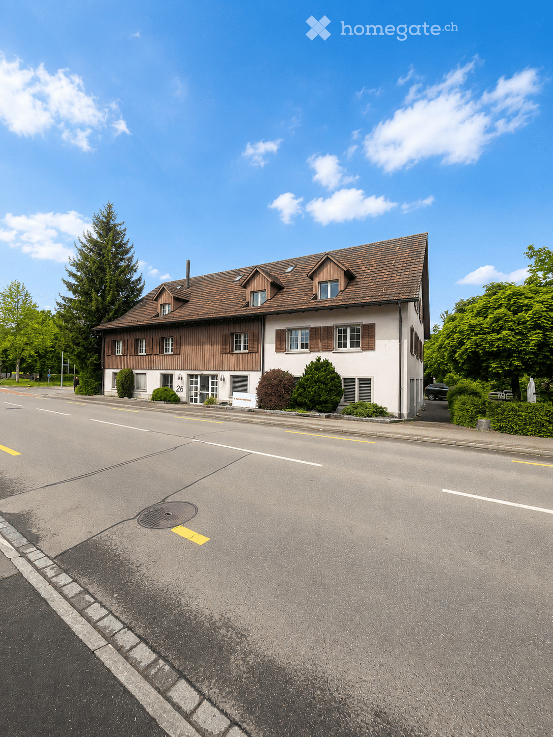 Two-story house with a brown tiled roof, white walls, and multiple windows with shutters. The house is surrounded by greenery and is located on a quiet street.