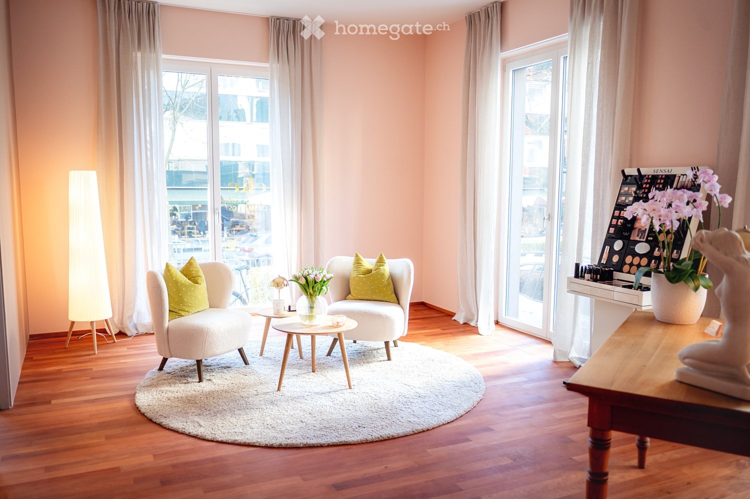 Furnished Room. Bright living room with wooden flooring, large windows, white sofa, coffee table, decorative pillows, and a display of cosmetics and flowers.