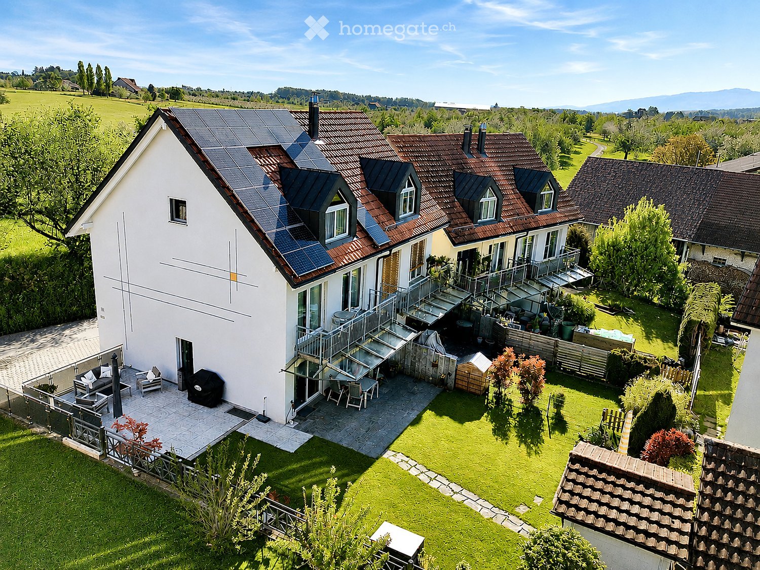 A modern two-story house with solar panels on the roof, surrounded by a well-maintained garden and outdoor seating area.