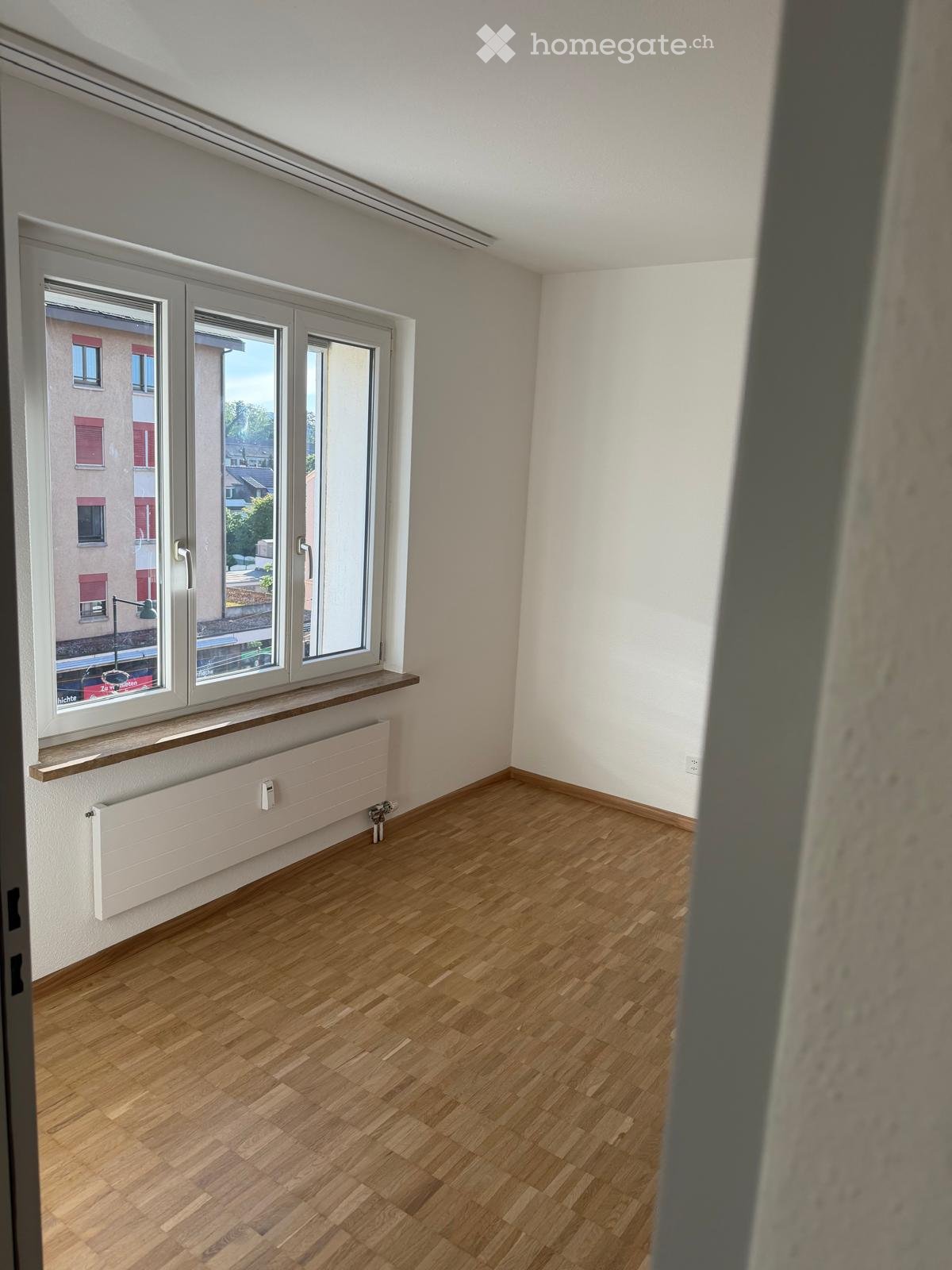 Unfurnished Room. Empty room with wooden flooring, white walls, large windows with a view of the street, and a radiator below the windows.
