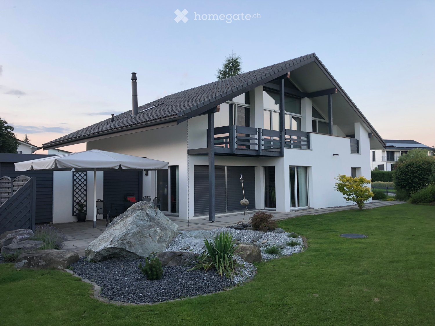 House. Modern two-story house with white exterior, large windows, and a balcony on the upper level. The front yard features a well-maintained lawn, decorative rocks, and a patio area with outdoor furniture.