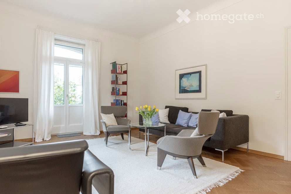 modern living room with white walls, wooden floor, white rug, gray couch, glass table, chairs, TV, bookshelf, picture frame, flowers