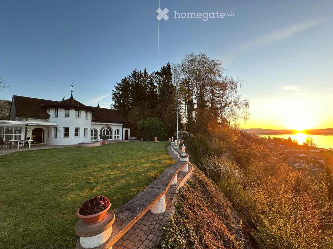 House. A large, white house with a dome-shaped roof and multiple windows, situated on a grassy lawn overlooking a body of water at sunset.