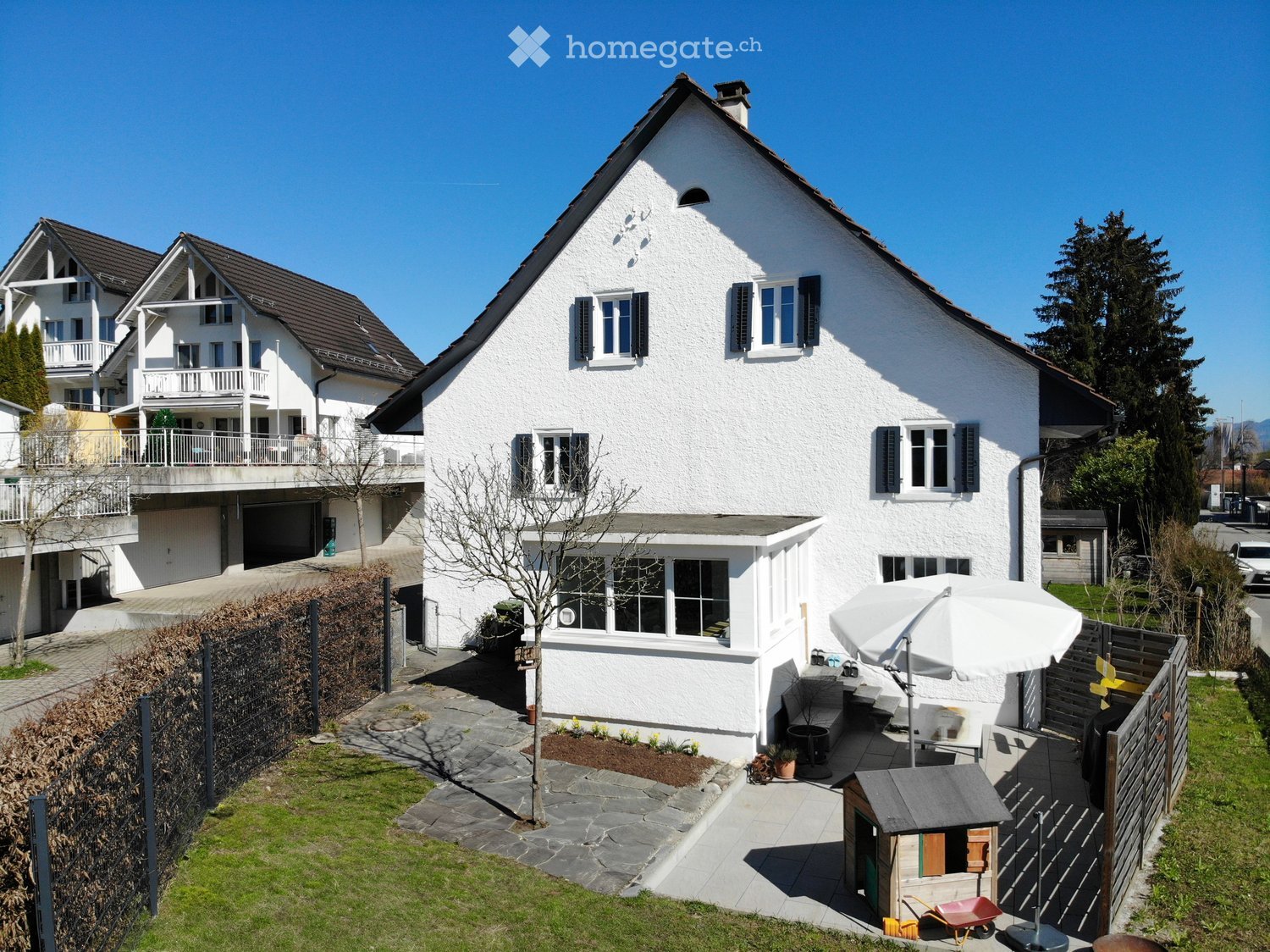 Two story house, white walls, brown roof, multiple windows, garden, patio, shed