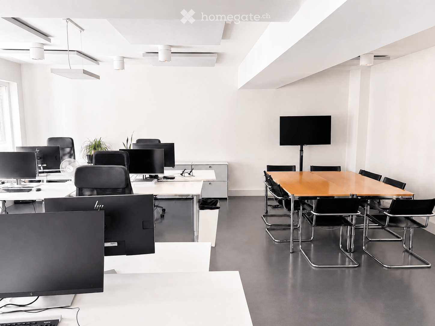 Modern office space with multiple workstations, a conference table, and a large screen. The room features sleek black chairs, desks, and a potted plant, with a polished dark floor and white walls.