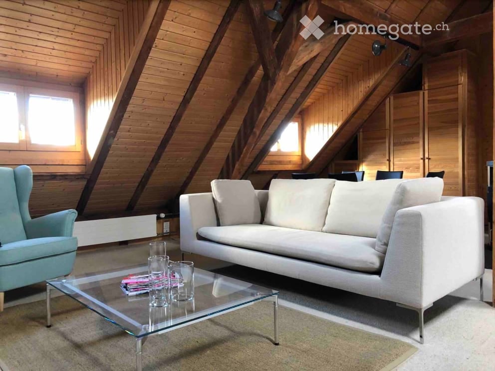 Living room with exposed wooden beams, large windows, a white sofa, a glass coffee table, and a light blue armchair.