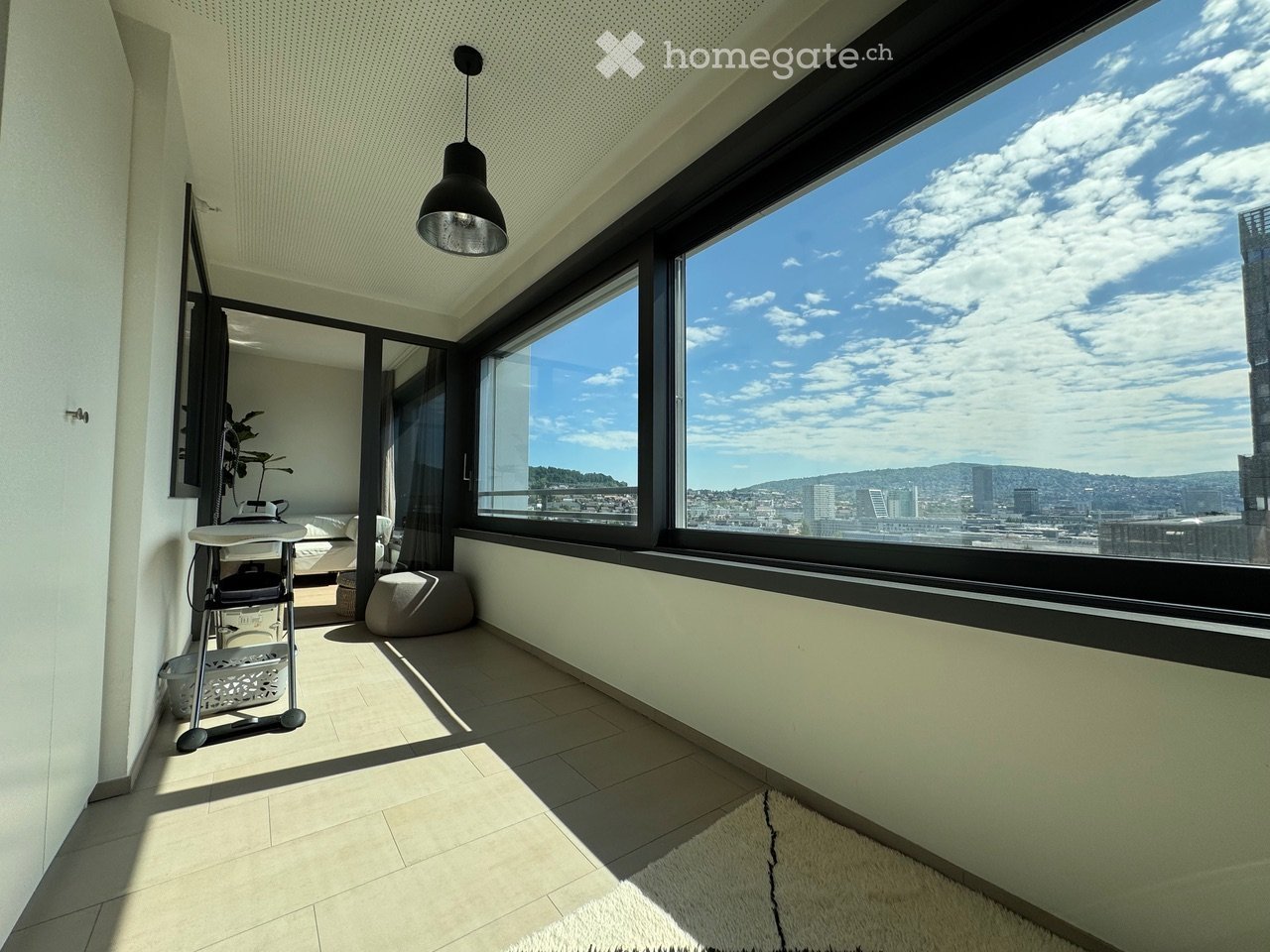 Balcony with large windows offering a city view, equipped with a small table, chair, and a round stool.