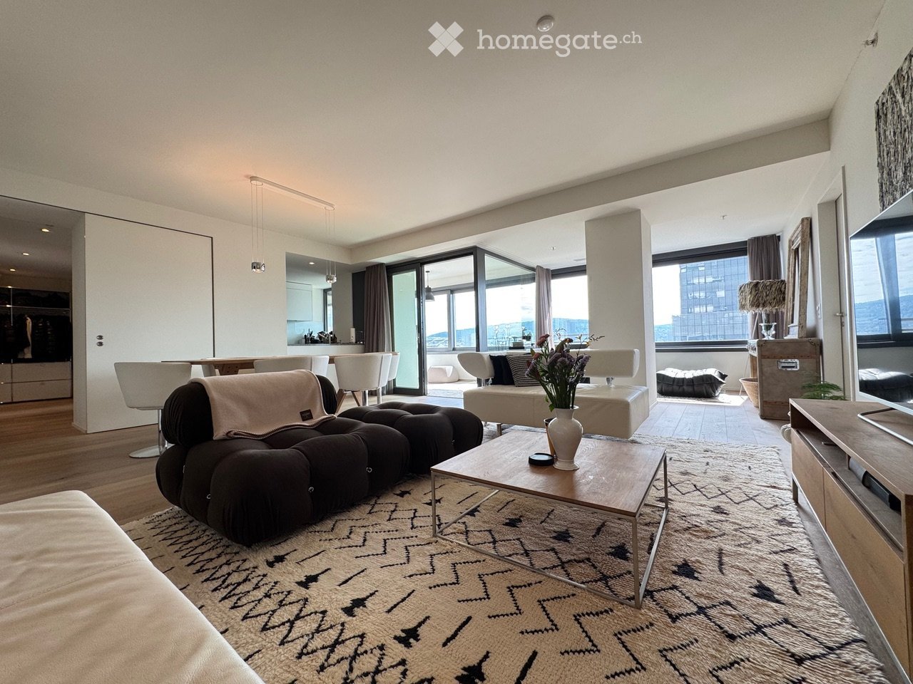Modern living room with large windows offering a city view, featuring a dark tufted sofa, coffee table, ottoman, and several chairs around a dining table.