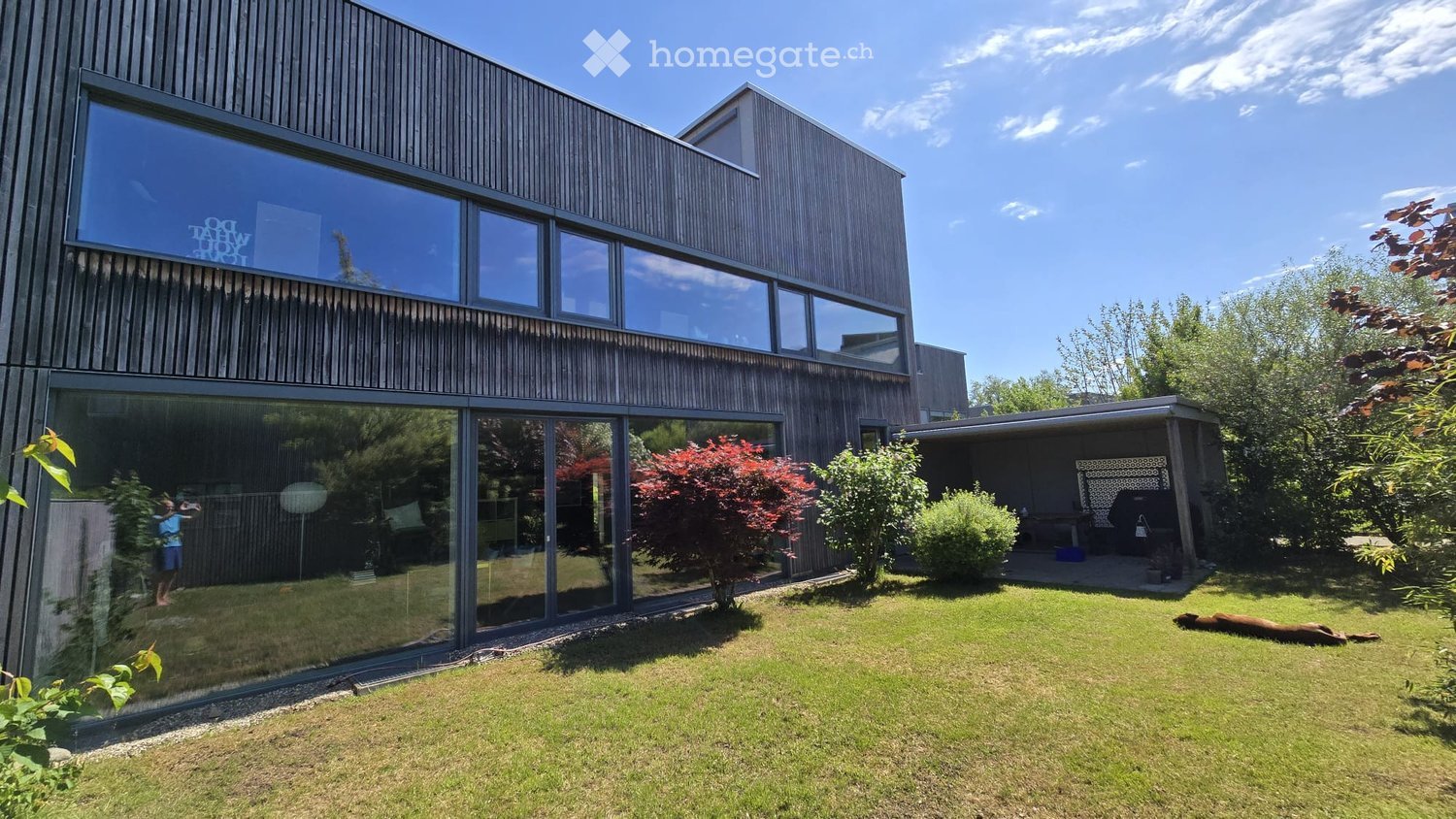 Garden / Outdoor Area. Modern house with large glass windows and wooden facade, surrounded by a well-maintained lawn, shrubs, and trees. A small outdoor seating area is visible to the right.