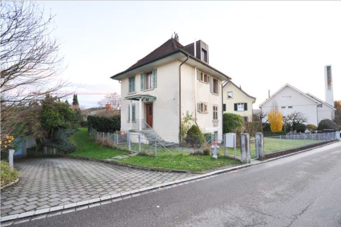 House. A two-story house with a cream-colored exterior, multiple windows, and a small porch. The house is surrounded by a well-maintained lawn and a low fence.