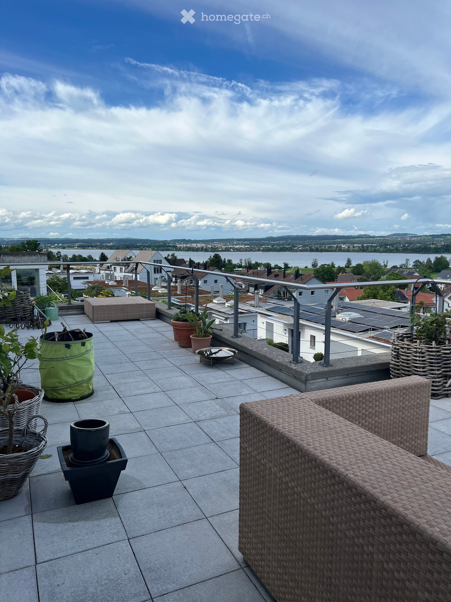 A spacious balcony with a tiled floor, a wicker sofa, several potted plants, and a scenic view of a city and lake in the background.