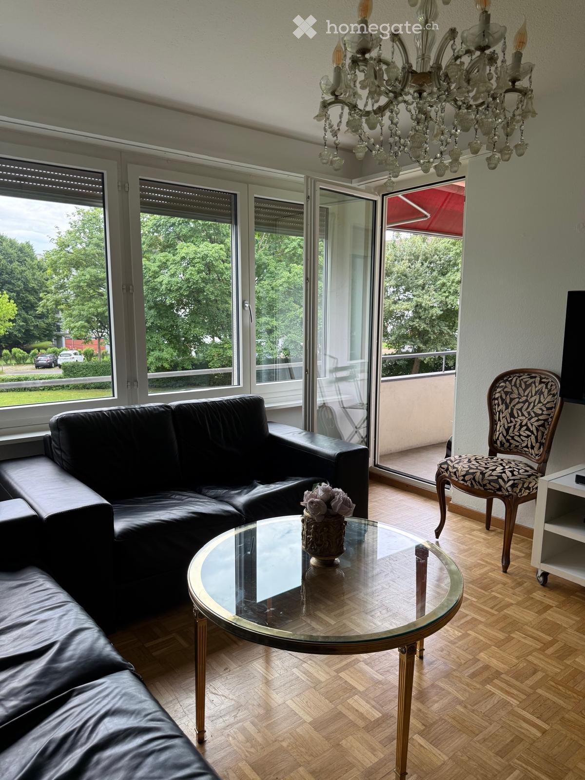 Modern living room with large windows and balcony access, featuring a black leather sofa, glass coffee table with floral centerpiece, patterned armchair, and ornate chandelier.