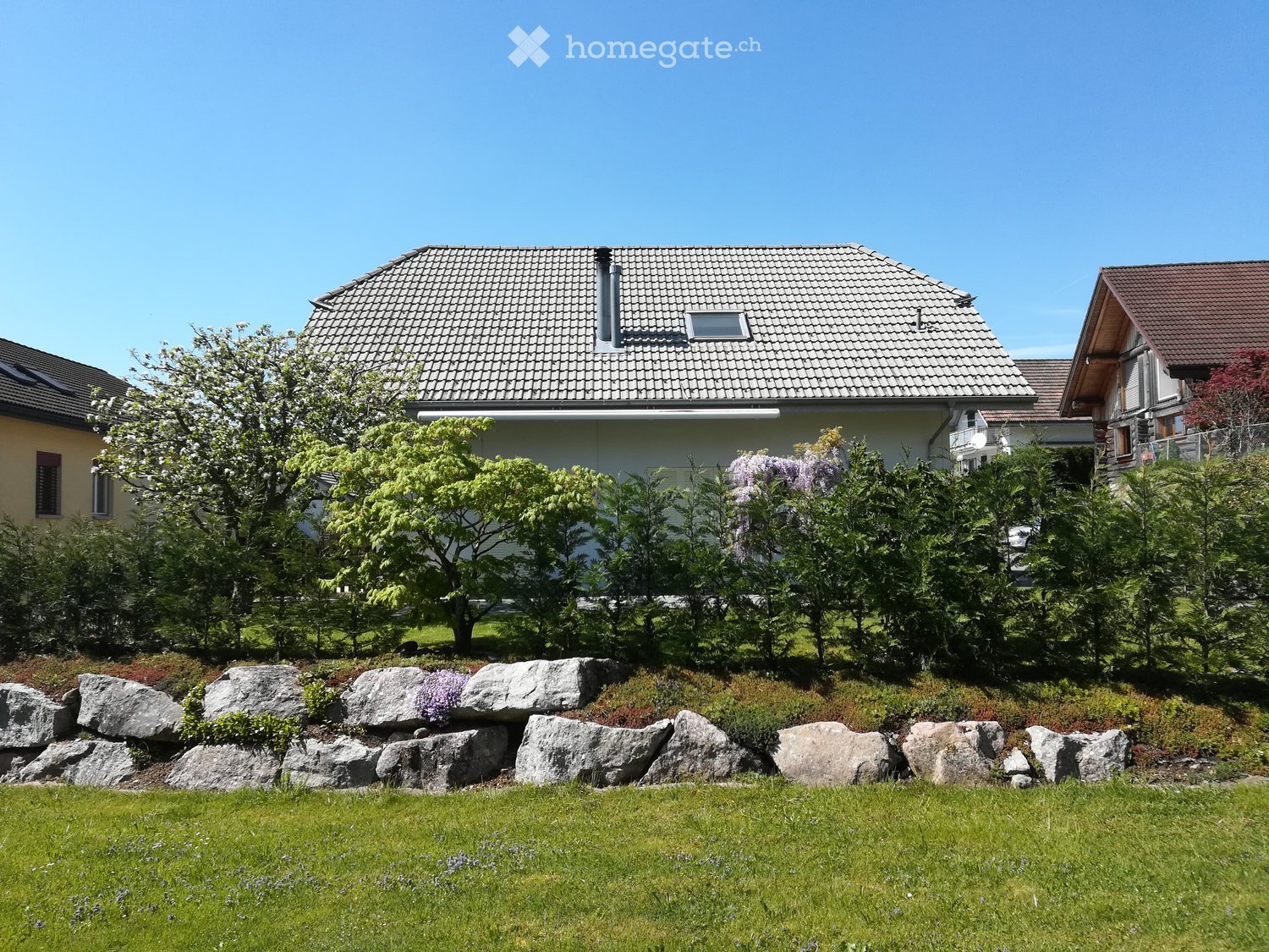House. Two-story house with a tiled roof, surrounded by a well-maintained garden with trees, shrubs, and a stone retaining wall.