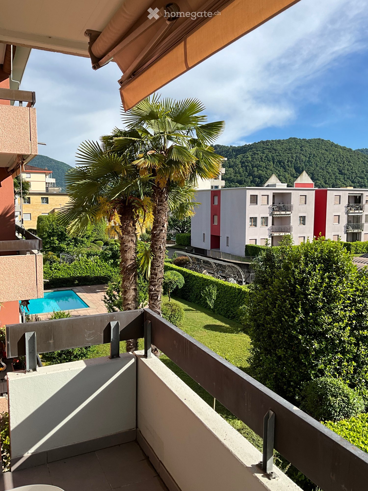 Balcony with a view of a landscaped garden, swimming pool, and surrounding buildings, featuring a palm tree and a mountainous backdrop.