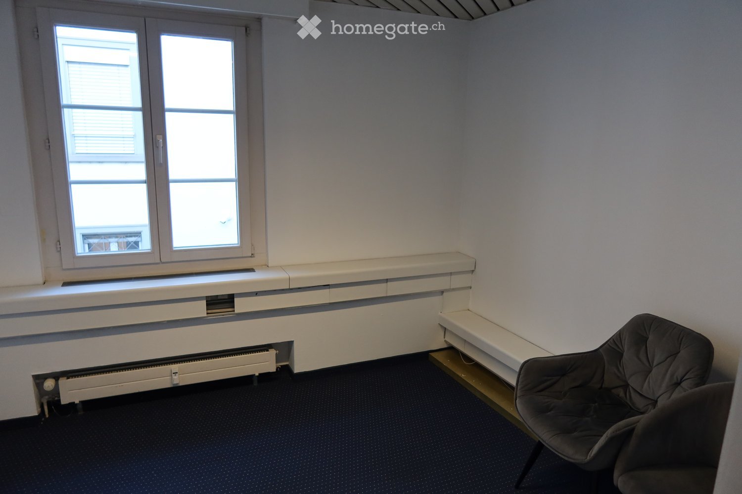 Empty room with white walls, white windowsill, window with shutters, bench, chairs, radiator, and dark carpet.