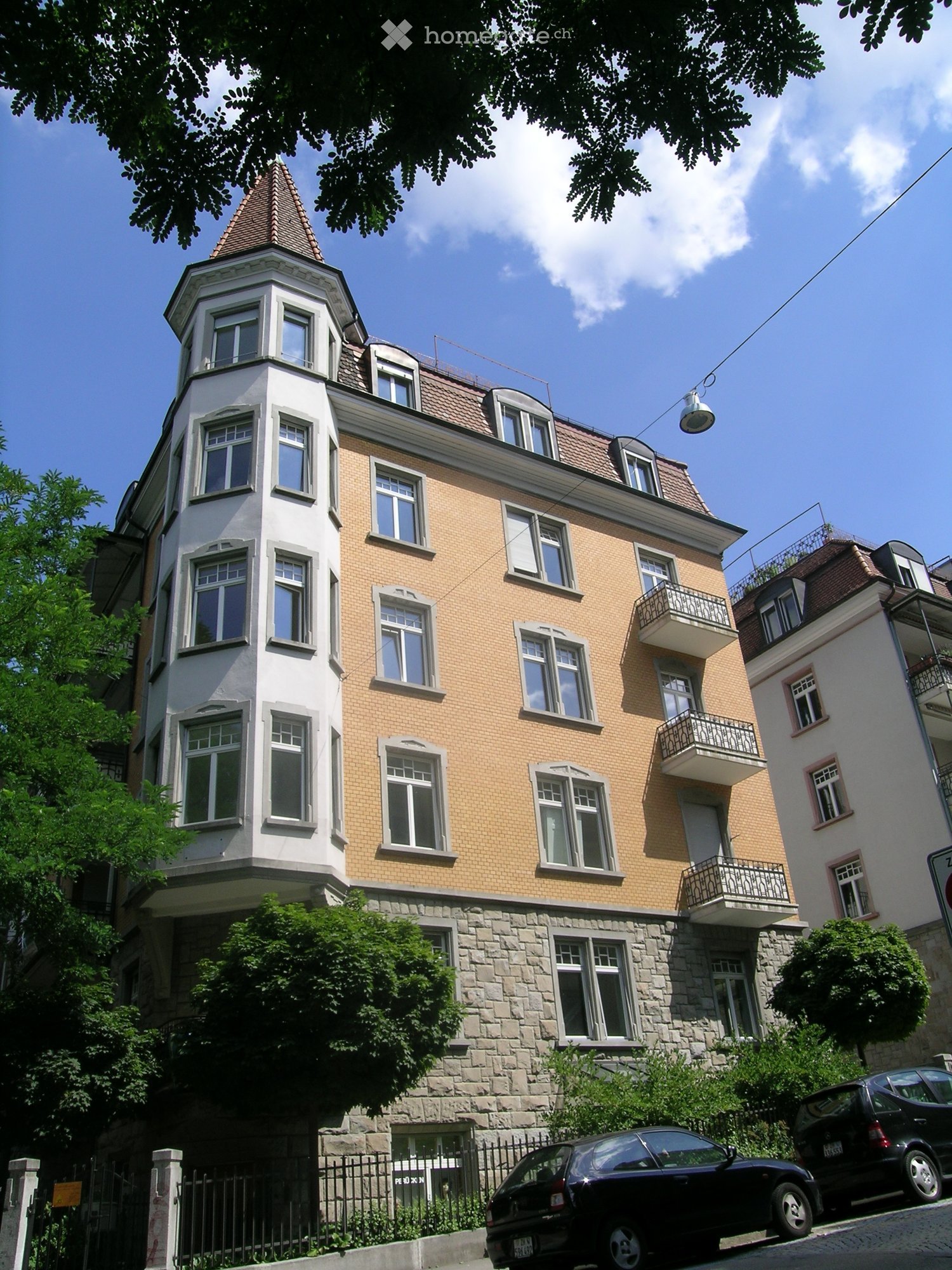 A multi-story residential building with a mix of brick and stone facades, multiple windows, and a prominent balcony on the upper floor. The building features a distinctive tower with a pointed roof and is surrounded by greenery and parked cars.