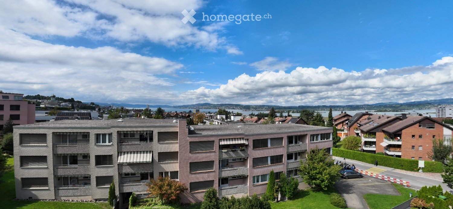 A multi-story building with multiple balconies, surrounded by greenery, with a view of the lake and mountains in the distance.